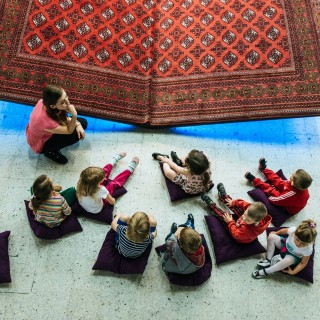 Jak ciekawie oprowadzać i pisać o dziedzictwie - szkolenie dla osób zajmujących się edukacją kulturową  i oprowadzaniem - Ciekawe opowiadanie wzbudza ciekawość i&nbsp;zaangażowanie. Na fotografii widać młodą dziewczynę opowiadającą dzieciom o&nbsp;zabytkowym dywanie powieszonym za jej plecami. Dzieci siedzą na poduszkach i&nbsp;słuchają.