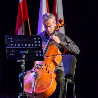Koncert Patriotyczny Kwartetu Smyczkowego Filharmonii Krakowskiej - Mężczyzna gra na wiolonczeli. W tle widać trzy flagi Polski