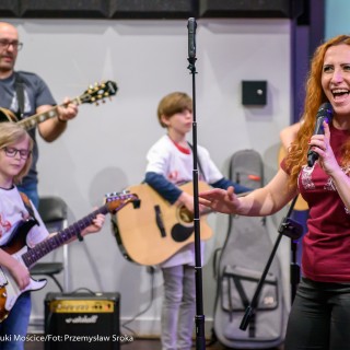 Akcja muzyczna na 50-lecie Centrum - Kobieta w bordowej bluzce śpiewa do mikrofonu, za nią w tle mężczyzna w okularach i dwóch chłopców gra na gitarach.  - Fot. : Przemysław Sroka